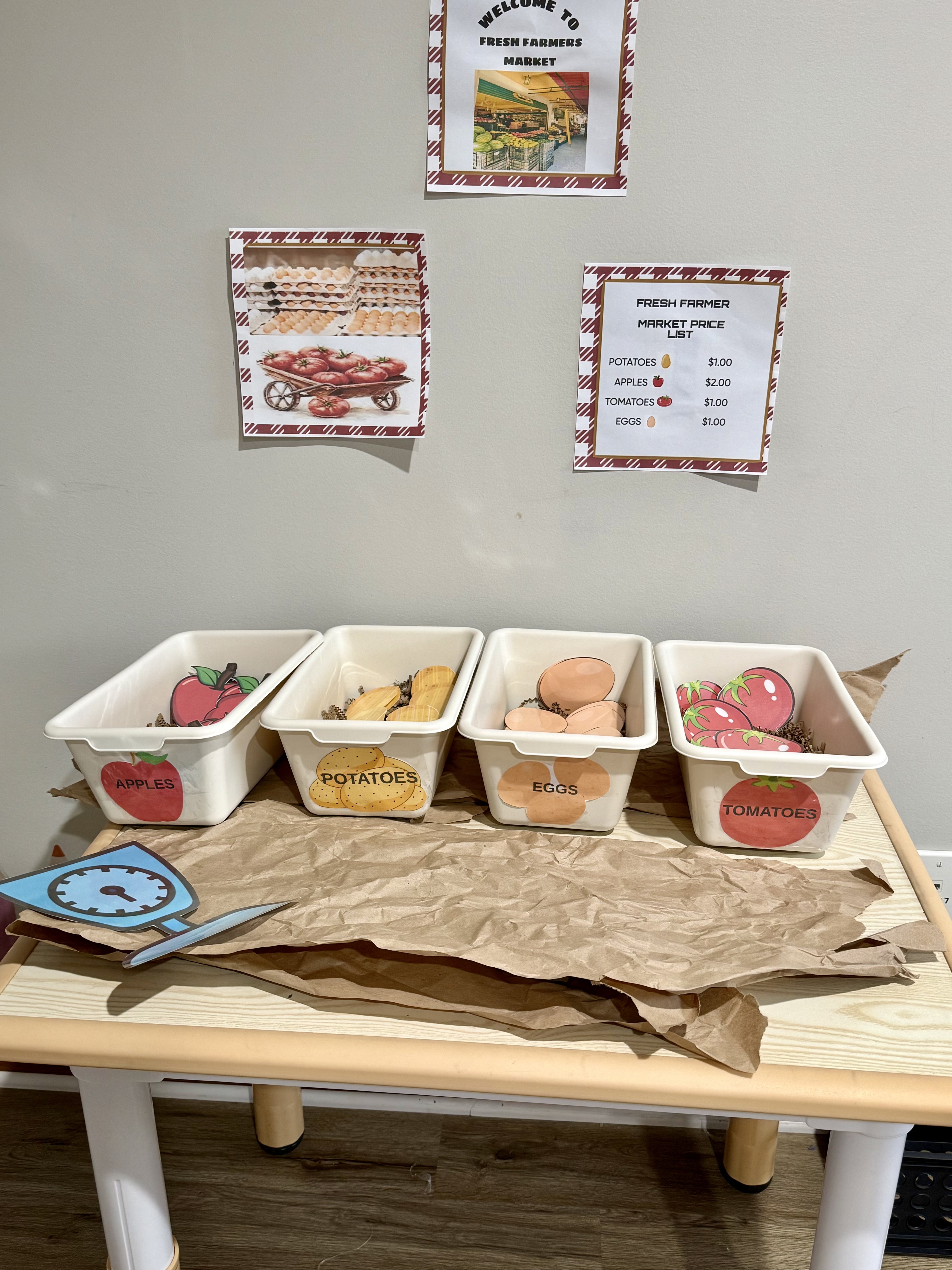 A pretend farmers market learning station with labeled bins for apples, potatoes, eggs, and tomatoes